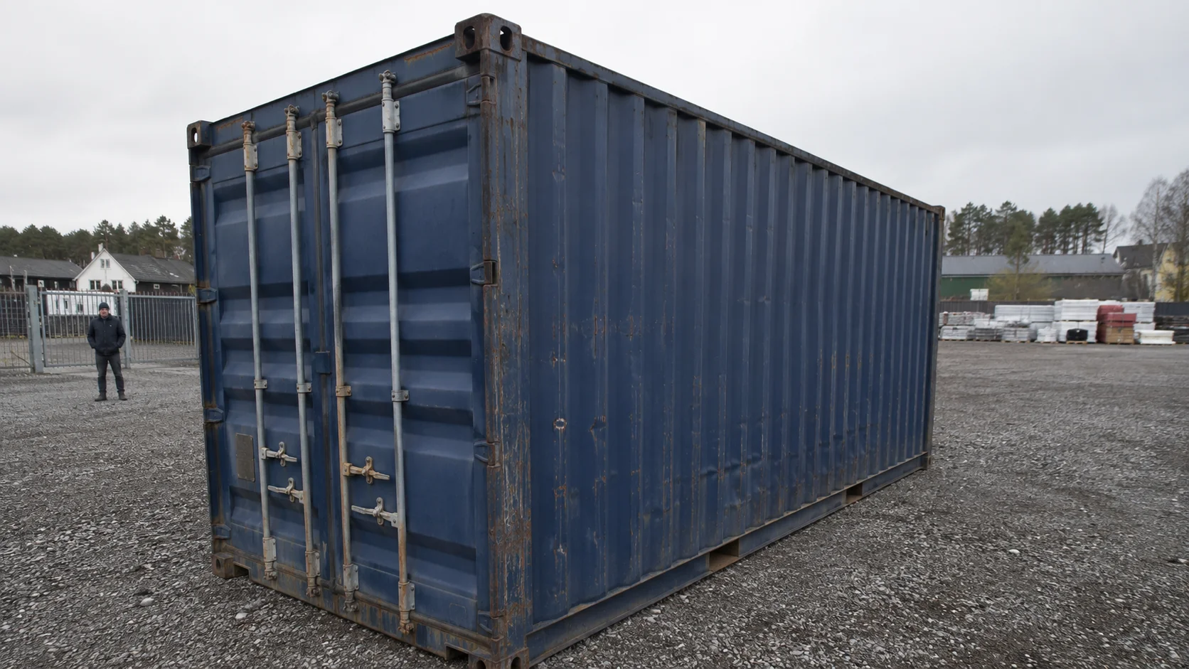 20 fods container set udefra på grus, med dørende synlig og målforhold til en person i baggrunden